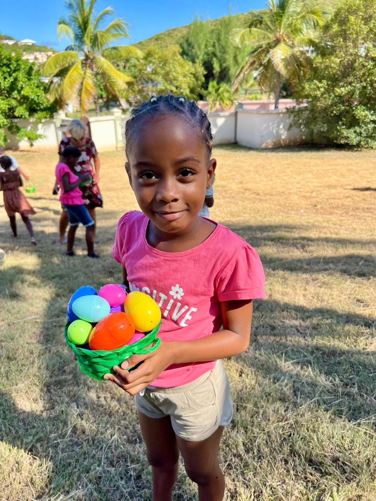 A child holding a green basket of colourful eggs outside, with palm trees in the background